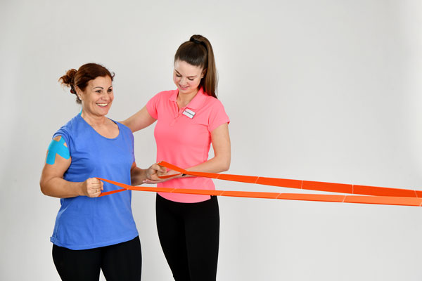 Eine Frau führt mit einem Trainingsband eine Schulterübung durch, unterstützt von einer Physiotherapeutin